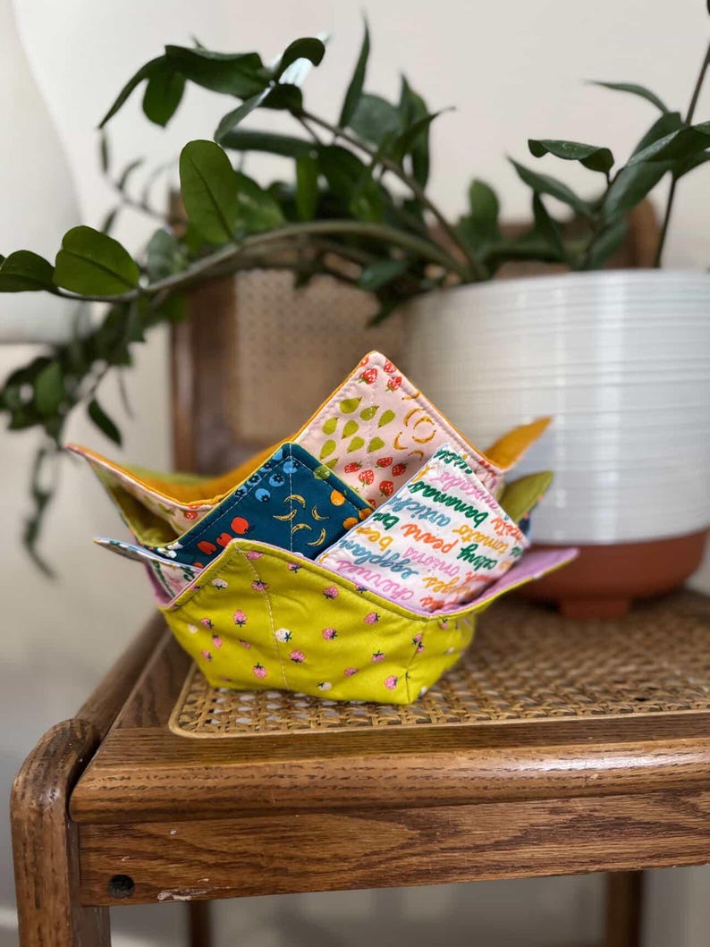 May include: A stack of colorful fabric bowls with various fruit and vegetable patterns. The bowls are yellow, pink, and teal, with designs of strawberries, bananas, and other produce. The bowls are stacked on a wooden table.