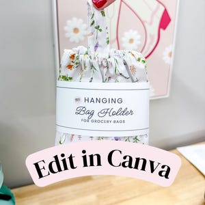 May include: A white fabric hanging bag holder for grocery bags featuring a colorful floral print of small wildflowers. The item is wrapped in a white paper band with the text HANGING Bag Holder FOR GROCERY BAGS printed in black ink.