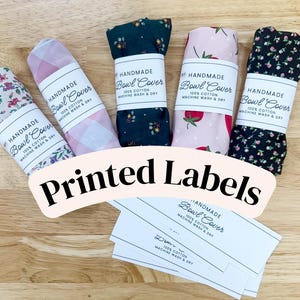 May include: Five folded fabric bowl covers with various patterns including floral, plaid, and strawberry prints. Each cover features a white paper band with the text Handmade Bowl Cover, 100% Cotton, Machine Wash & Dry. Several loose labels are included.