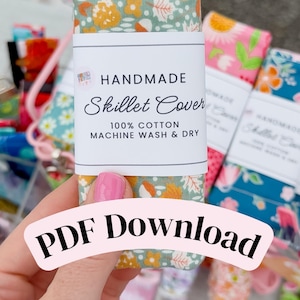 May include: A rectangular fabric skillet cover with a sage green background featuring a pattern of yellow flowers, white leaves, and orange acorns. A white paper label reads HANDMADE Skillet Cover 100% COTTON MACHINE WASH & DRY. PDF DOWNLOAD.