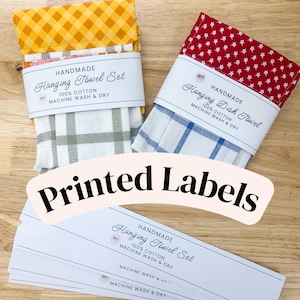 May include: Two folded hanging towels with paper bands. One towel has a yellow gingham top and white and sage green plaid bottom. The other has a red floral top and white and blue plaid bottom. Several white paper labels with black text are included.