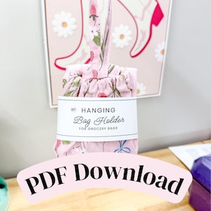Puede incluir: Un soporte de tela rosa claro para colgar bolsas de la compra con un estampado de fresas rojas, hojas verdes y flores blancas. Una etiqueta de papel blanco envuelve el centro con el texto HANGING Bag Holder FOR GROCERY BAGS.