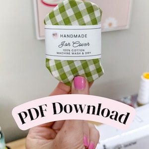 Puede incluir: Una tapa de tarro hecha a mano de tela 100% algodón con un estampado de cuadros verdes y blancos. La etiqueta dice "HANDMADE Jar Cover 100% COTTON MACHINE WASH & DRY".