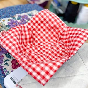 May include: A red and white gingham patterned bowl cozy. The square-shaped cozy is open and ready to hold a bowl. The cozy is sitting on a white surface with a floral patterned fabric in the background.