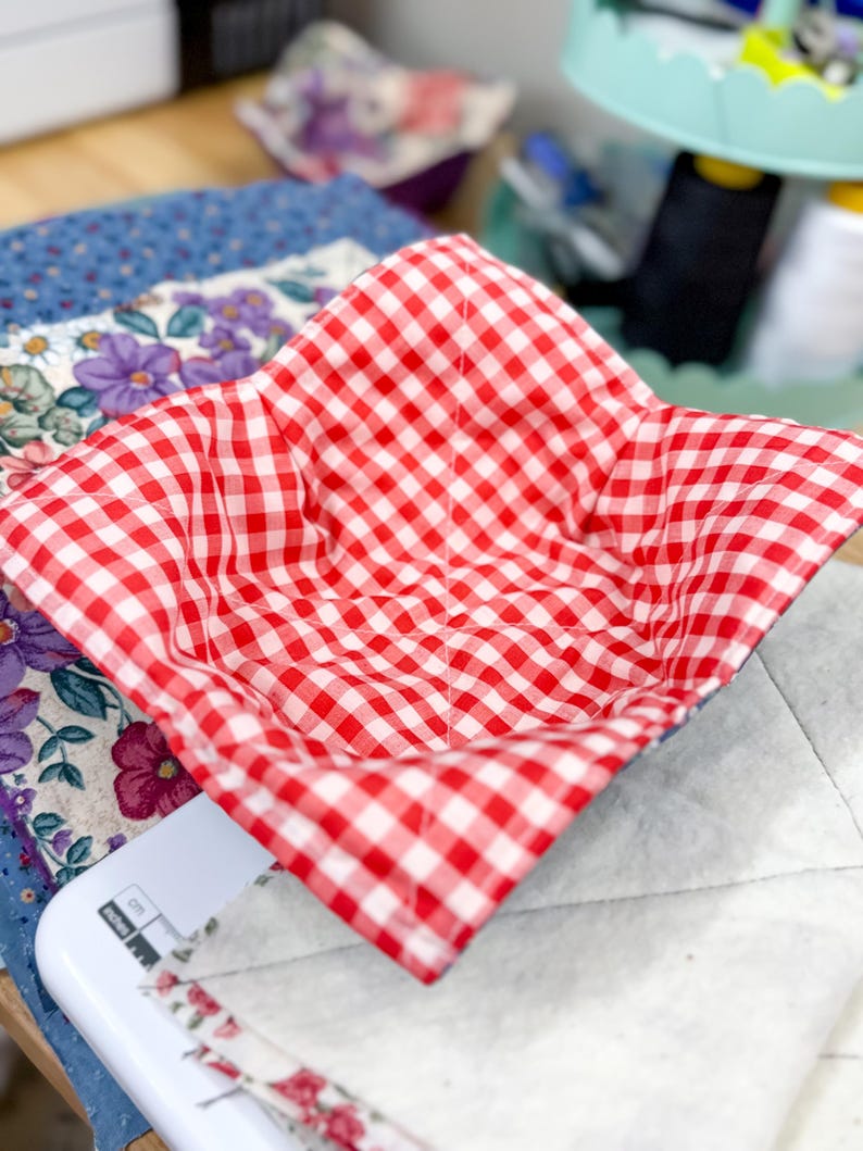 May include: A red and white gingham patterned bowl cozy. The square-shaped cozy is open and ready to hold a bowl. The cozy is sitting on a white surface with a floral patterned fabric in the background.