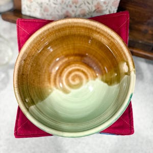 May include: A handmade ceramic bowl with a brown and green glaze, resting on a red quilted square. The bowl's interior features a spiral design. The background includes a floral-patterned package and a wooden shelf.