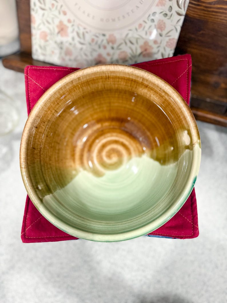 May include: A handmade ceramic bowl with a brown and green glaze, resting on a red quilted square. The bowl's interior features a spiral design. The background includes a floral-patterned package and a wooden shelf.