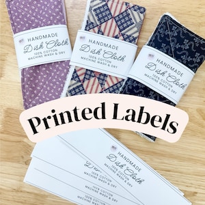May include: Three folded dish cloths with purple, plaid, and dark blue floral patterns, each wrapped in a white paper band labeled HANDMADE Dish Cloth 100% COTTON MACHINE WASH & DRY, alongside a stack of identical blank paper labels.