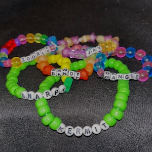May include: A collection of colorful beaded bracelets. The bracelets are made of various colored beads, including green, yellow, orange, pink, blue, and purple. Some bracelets have white letter beads spelling out names like "ZOOBLE", "DAN DY", "SNARPY", "KERMIT", "G GANGLE", and "JAX".