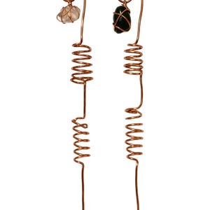 May include: Two copper-colored plant stakes with spiral designs. One stake has a clear, irregularly shaped stone at the top, while the other has a dark green stone. The stakes are designed to be inserted into the soil.