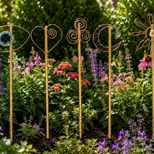 May include: Five decorative garden stakes made of gold-toned wire, each featuring a unique top design: an eye with a blue stone center, a heart with a spiral, a triple spiral, a crescent moon with a clear crystal, and a sun with a yellow stone.