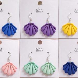 May include: A selection of seashell earrings in shades of blue, purple, yellow, pink, mint green, and navy. Each pair of earrings is presented on a white card with a silver hook. These earrings are a bright and colourful accessory.
