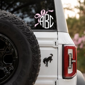 May include: White monogram decal with the letters "ABC" inside a circle, adorned with a pink bow and ribbons. The decal is affixed to the rear window of a white vehicle. A black spare tire is visible on the left.