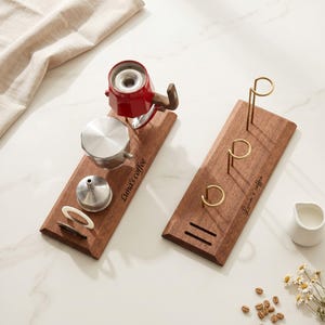 May include: Two wooden coffee station organisers with metal and ceramic coffee brewing equipment. One organiser holds a red coffee maker, a silver filter, and other accessories. The other has gold-coloured holders. The text "Luna's coffee" is on each.
