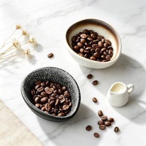 Può includere: Due piccole ciotole in ceramica asimmetriche piene di chicchi di caffè tostati. Una ciotola è nera con un motivo maculato, l'altra è bianca con un bordo marrone. Un piccolo bricco per panna bianco e chicchi di caffè sparsi completano la composizione.