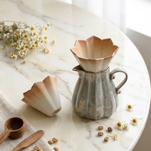 May include: A ceramic coffee pour-over set with a flower-shaped filter and a ribbed pitcher. The filter is light brown and white, and the pitcher is gray. The set is on a marble table with coffee beans and flowers.