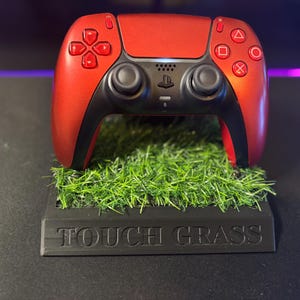 May include: A metallic red video game controller resting on a black rectangular stand featuring a patch of artificial green grass on top and the text TOUCH GRASS embossed in capital letters on the front face of the base.