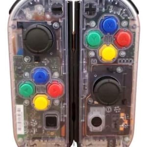 May include: Clear, transparent video game controller with visible internal components. Features colored buttons in blue, green, red, and yellow, along with black joysticks and directional pads. The controller is designed for a gaming console.