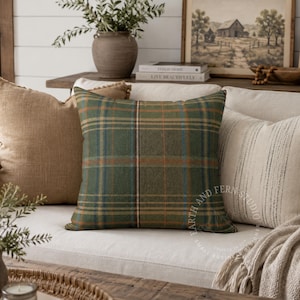 May include: A square throw pillow featuring a green, brown, and blue plaid pattern. The fabric has a textured appearance with intersecting horizontal and vertical lines. The pillow is positioned centrally among other neutral-colored cushions.