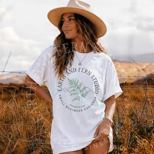 Könnte beinhalten: Weißes T-Shirt mit einem kreisförmigen Grafikdesign mit dem Text "Earth and Fern Studio" um ein Farn- und Bergdesign. Das Shirt enthält auch den Text "Small Business Designer" und "Shipped in USA".