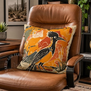 May include: A square throw pillow featuring a graphic illustration of a roadrunner bird in black against a vibrant background of orange, rust, and muted green desert shapes. The design includes stylized cacti and abstract textures on a cream base.
