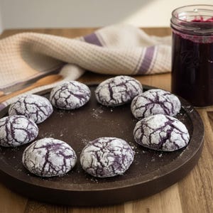 May include: A circular arrangement of eight purple crinkle cookies dusted with powdered sugar on a dark brown plate. A jar of purple jam sits to the right. A light-colored cloth and wooden spoon are in the background.