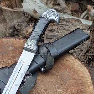 Può includere: Un pugnale argento e nero con un'impugnatura a testa d'aquila e un fodero in pelle. La lama lucida è riflettente. Il pugnale è appoggiato su una superficie di legno. Il testo "MONARCH ARMOURY" è visibile.