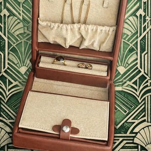 May include: Open brown leather jewelry box with multiple compartments. The top section holds gold necklaces, while the middle section displays rings. The bottom section is closed with a leather strap and a silver button. The interior is beige.