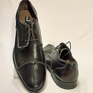 May include: A pair of dark brown leather oxford shoes featuring a cap toe design with a vertical seam down the center, dark laces, and a visible brand label on the inner heel. The shoes have a polished finish and a low stacked heel.