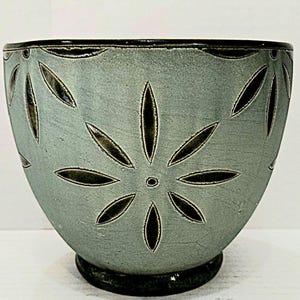 May include: A teal ceramic bowl with a dark rim and base. The bowl features a floral design with dark, leaf-shaped cutouts. The bowl has a textured surface and a rounded shape, suitable for serving or display.