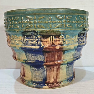 May include: A handcrafted ceramic planter with a turquoise top, featuring embossed floral patterns. The planter transitions to blue, brown, and beige vertical stripes. A unique, decorative piece for indoor or outdoor use.