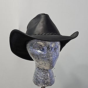 Peut inclure: Un chapeau de cowboy noir avec un large bord incurvé et une bande en cuir tressé autour de la base de la calotte. Le chapeau présente un design structuré avec un sommet pincé et est présenté sur un mannequin de tête texturé argenté.