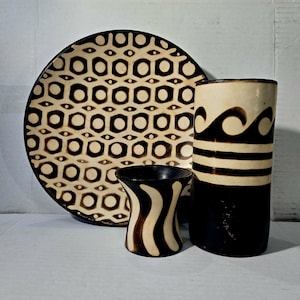 Puede incluir: Un conjunto de tres piezas de cerámica hechas a mano: un plato grande con un patrón geométrico, un jarrón alto con diseños de olas y rayas, y una taza pequeña con un patrón de rayas onduladas. Las piezas son de color marrón y crema.