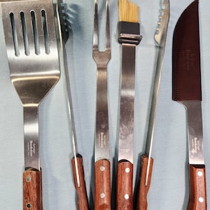 May include: A set of five stainless steel grilling tools with wooden handles, including a slotted spatula, tongs, a two-pronged fork, a basting brush with tan bristles, and a knife with a brown leather sheath, all marked Heritage Brookstone.