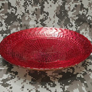 May include: A textured, oval bowl in a striking red colour. The bowl's surface features a woven design, and the edges are slightly raised. The bowl is likely made of glass or a similar material. The background has a camouflage pattern.