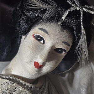 May include: A close-up of a Japanese doll with a pale face, black hair, and red lips. The doll has detailed facial features, including dark eyes and delicate eyebrows. Feathers and a silver ribbon adorn the hair, and the doll is wearing a white kimono.