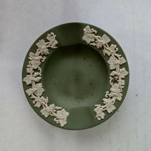 May include: A round, olive-green ashtray with a raised, cream-colored grape vine design around the rim. The ashtray has a smooth, matte finish and a small indentation for resting cigarettes.