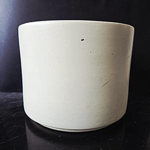 May include: A cylindrical, off-white ceramic planter with a slightly rounded base. The planter has a smooth, matte finish and shows minor imperfections. The interior is not visible. The planter is suitable for indoor or outdoor use.