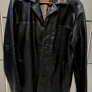 May include: A black leather coat with a notched collar and a button closure. The coat has a classic design with a tailored fit, and is hanging on a wooden hanger. The coat has two front pockets and a dark brown interior lining.