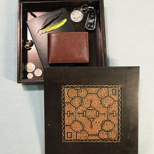 May include: A dark brown wooden valet tray with a square lid featuring an orange and black geometric pattern. Inside are a brown leather wallet, a black notebook, two feathers, a silver keychain, a car key fob, and three silver coins.