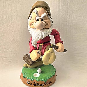 May include: A Grumpy figurine from Snow White and the Seven Dwarfs, wearing a brown hat and red tunic, holding a golf club over a green base with a golf ball and hole. The base features the text Walt Disney World in black script lettering.