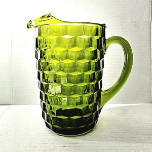 May include: A green glass pitcher with a geometric cube pattern. The pitcher has a curved handle and a spout for pouring. The pitcher is made of a transparent green glass.