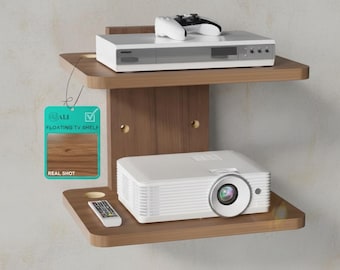 May include: A two-tier brown wooden floating shelf with rounded corners. The top shelf holds a white media player and a game controller. The bottom shelf holds a white projector and a remote. A teal tag reads ALI FLOATING TV SHELF REAL SHOT.