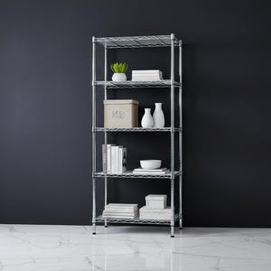 May include: A five-tier chrome wire shelving unit against a dark gray wall. The shelves hold a small potted plant, books, boxes, white vases, and a white bowl. The floor is white marble.