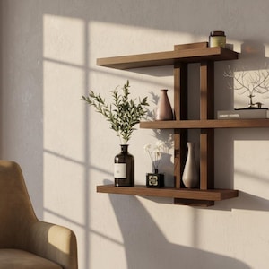 May include: A modern, dark wood wall shelf with three levels, holding decorative items. The shelf features a geometric design and displays vases, a small book, a candle, and a decorative tree sculpture. A vase of greenery and a small bottle are also visible.