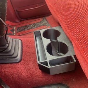 May include: Black plastic car cup holder with two circular cup slots and two rectangular slots. The cup holder is placed on a red carpeted floor next to a red car seat. The floor mat has the word "RACING" on it.