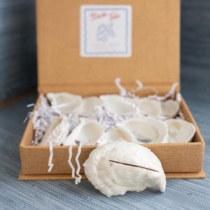 May include: A brown box filled with white oyster shells, some with a cut. The box has a label that says "Thank You". The shells are nestled in white paper. The box is on a blue surface.