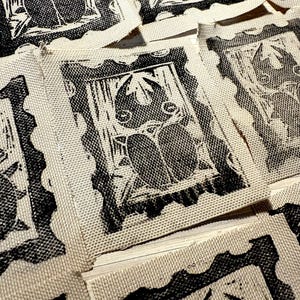 May include: Close-up of several fabric patches with a black and white linocut design. Each patch features a rectangular frame with a stylized image of a creature. The patches are made of a light-colored fabric with a woven texture.