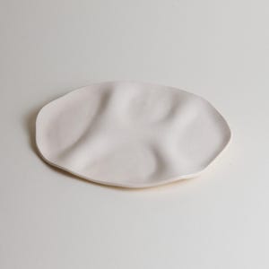 May include: A pale, off-white ceramic plate with an organic, undulating shape. The plate has a smooth surface and a slightly irregular edge, suggesting a handmade quality. The design is minimalist and modern, suitable for serving food or as a decorative piece.