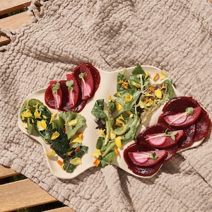 May include: A beige, organically shaped plate with a variety of colorful food items. The plate contains sliced beets, green leafy vegetables, and yellow flower petals. The plate sits on a beige textured cloth on a wooden table.